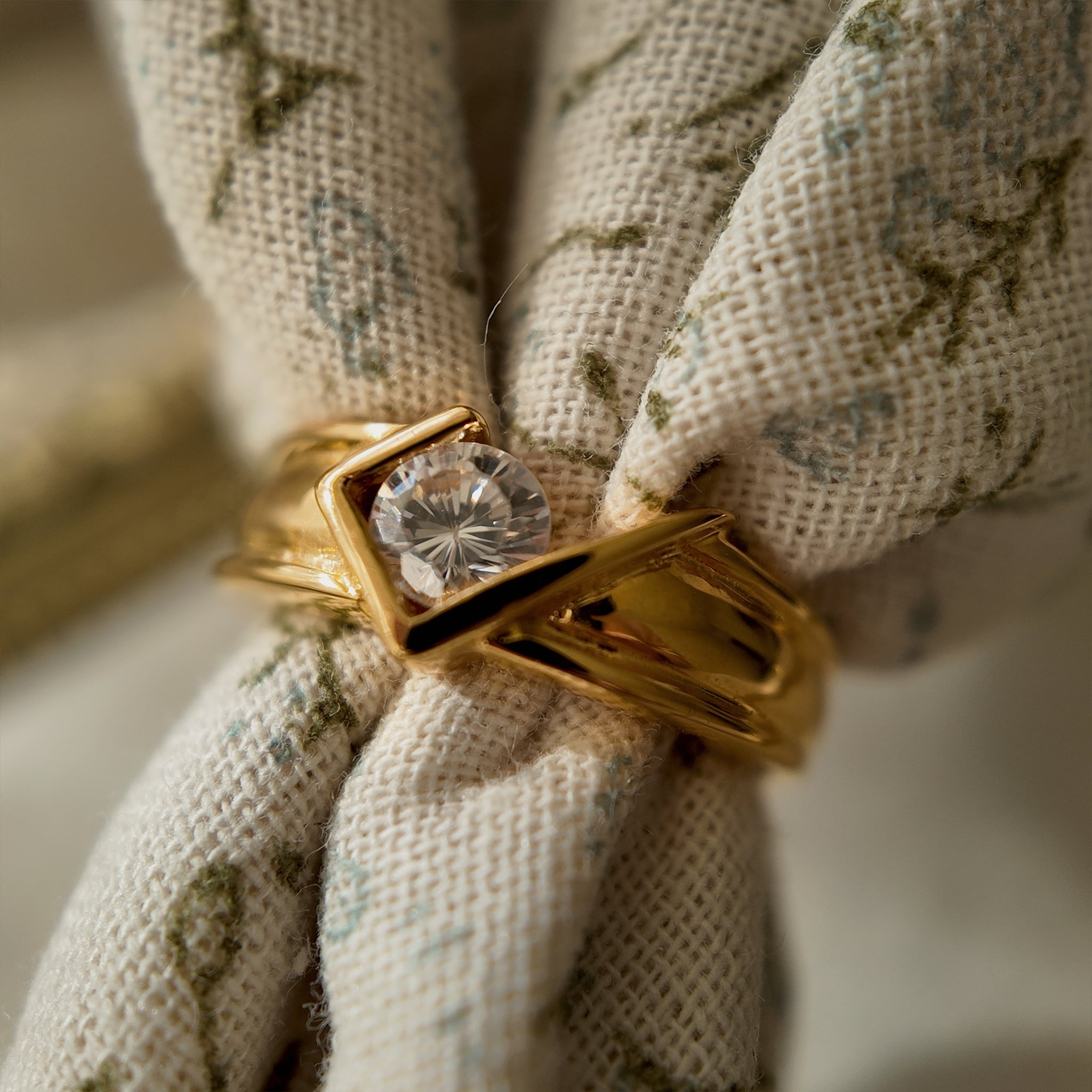 This 23k gold-plated ring features a square band set with round glass diamonds, while the band itself is plain gold. It boasts a simple geometric design. A piece of floral fabric is threaded through the ring to simulate how it would look on a hand.