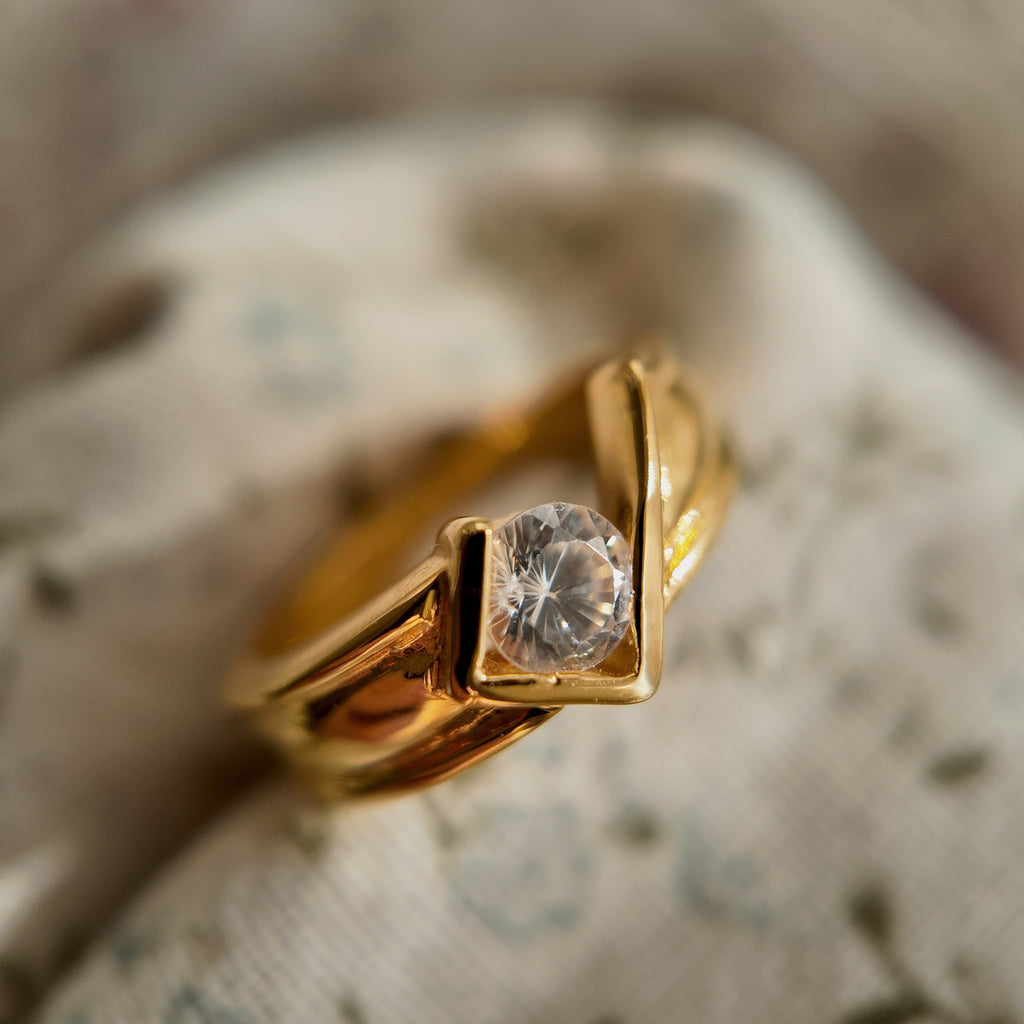 This 23k gold-plated ring features a square band with a round glass diamond set in it. The band itself is plain gold with a simple geometric design. When held up, it showcases the multifaceted facets and brilliance of the glass diamond.