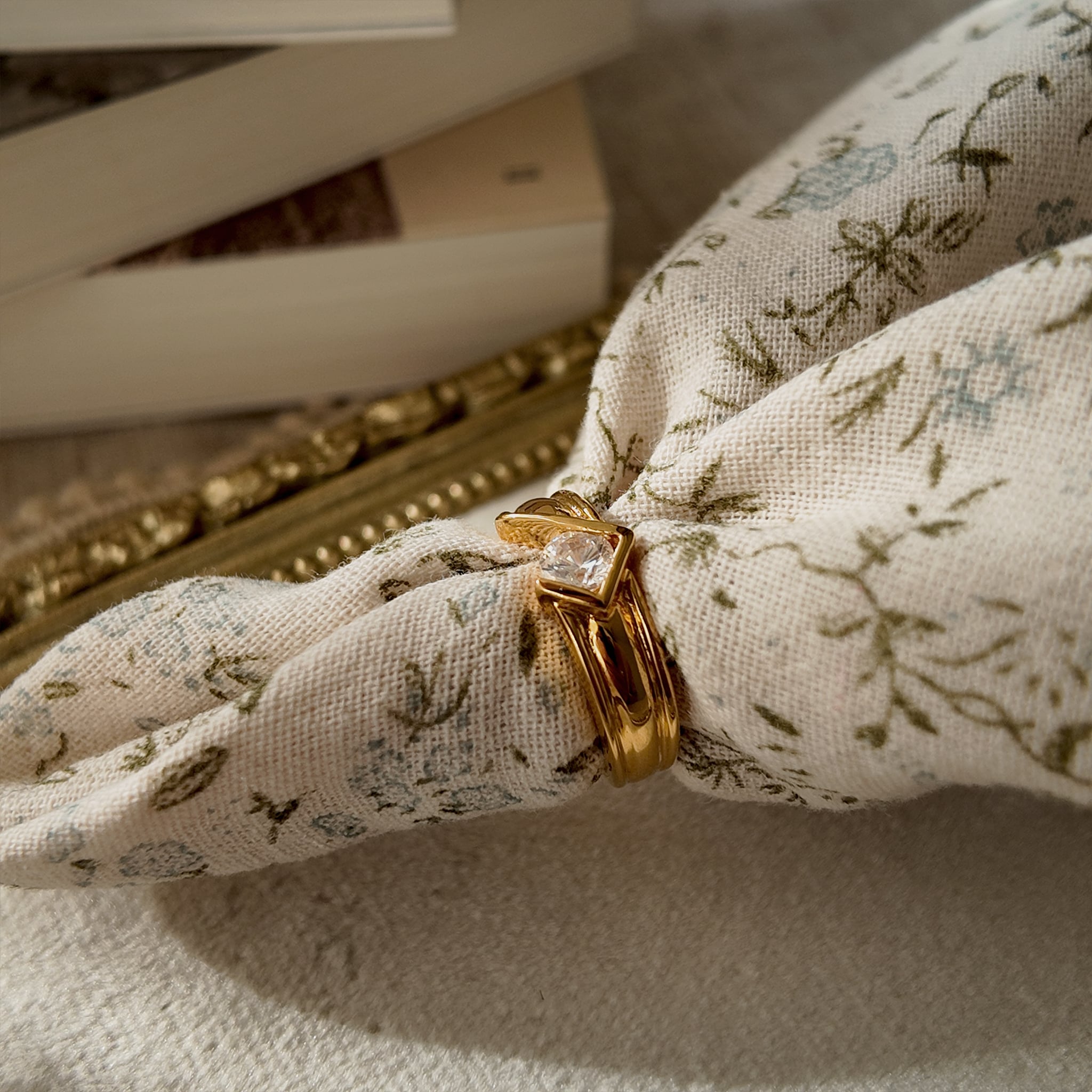 This 23k gold-plated ring features a square band set with round glass diamonds, while the band itself is plain gold with a simple geometric design. The side view, simulating how the ring is worn, showcases the integrity of the plating.