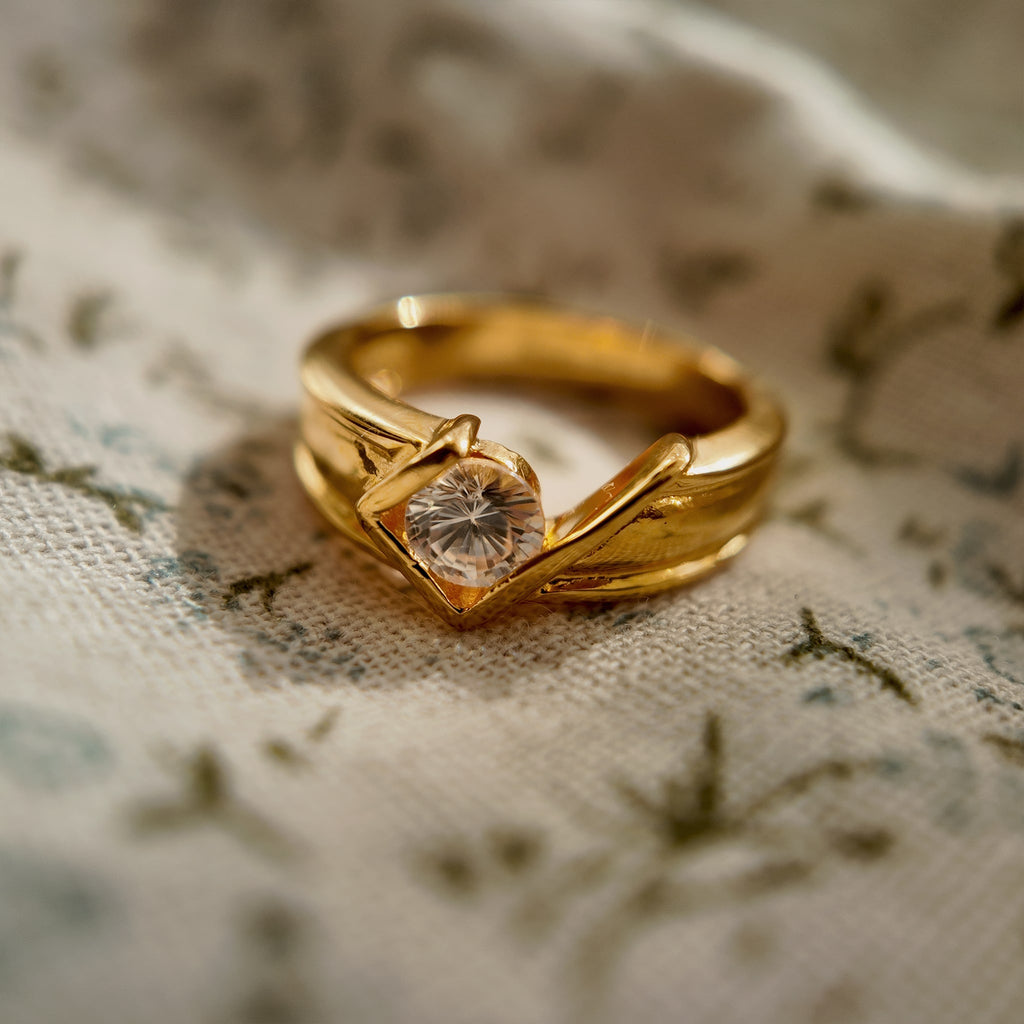 This 23k gold-plated ring features a square setting with round glass diamonds, while the band itself is plain gold with a simple geometric design. The image shows the setting and diamond arrangement from a different angle.