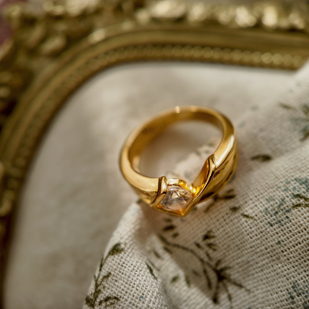 This 23k gold-plated ring features a square band set with round glass diamonds, while the band itself is plain gold with a simple geometric design. The image shows the side of the ring where the glass diamonds meet the band.