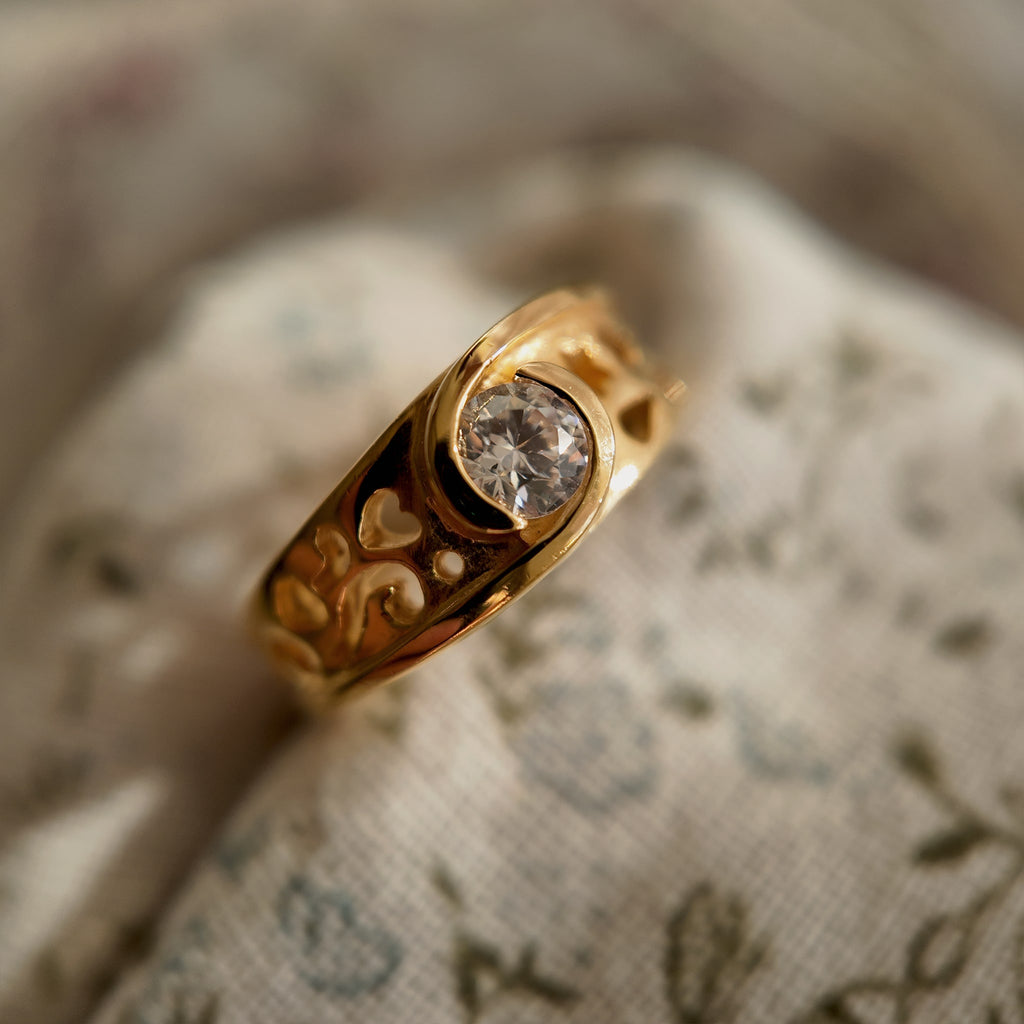 A 23k gold-plated ring features a round glass diamond, and the band is openworked with intricate patterns. A vintage ring set with a round glass diamond is shown being lifted to reveal the details on the front.