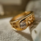 A 23k gold-plated ring features a round glass diamond, with the band openwork engraved with intricate patterns. The vintage ring is displayed suspended in a velvet case, allowing for a magnified view of the glass diamond's details.