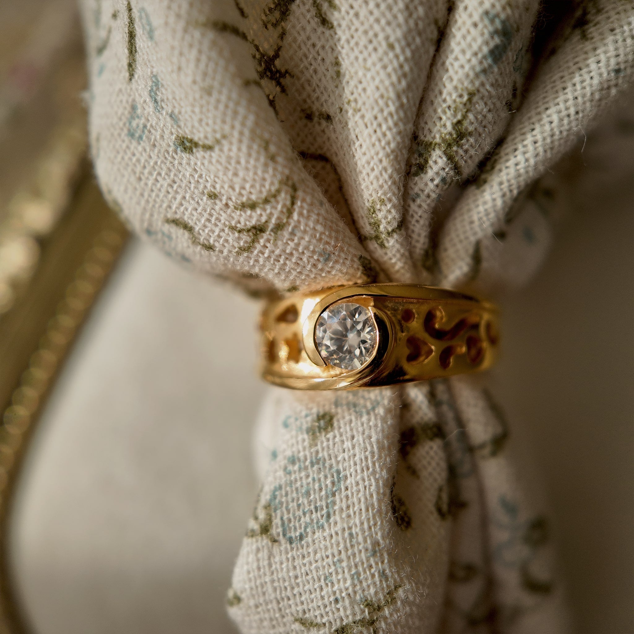 A 23k gold-plated ring features a round glass diamond set in a single piece, with the band openwork engraved with intricate patterns. The vintage ring is shown through a floral fabric weave, and a magnified view of the glass crystal reveals the different facets of the glass diamond.
