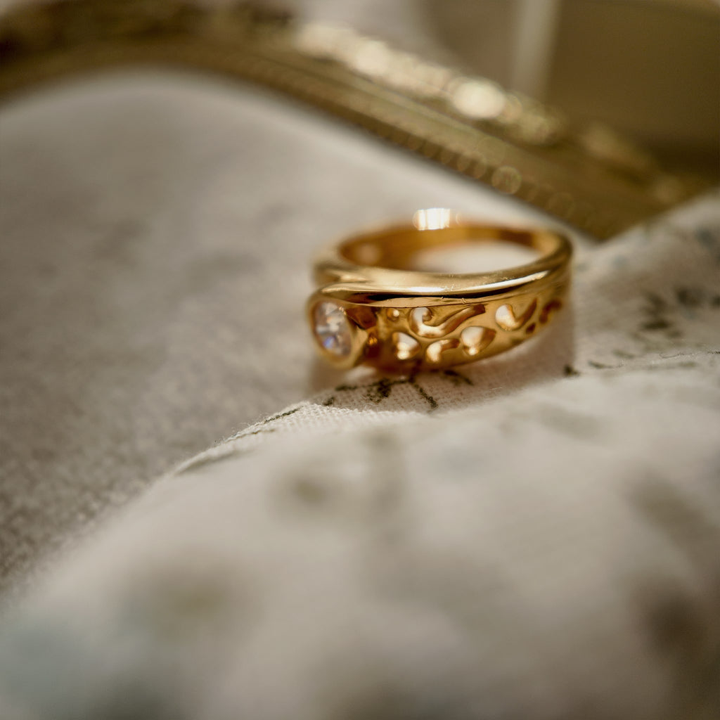 A 23k gold-plated ring is set with a round glass diamond, and the band is openwork-carved with intricate patterns. The vintage ring is displayed suspended in a velvet frame, with a side view that magnifies the details of the engraved designs.