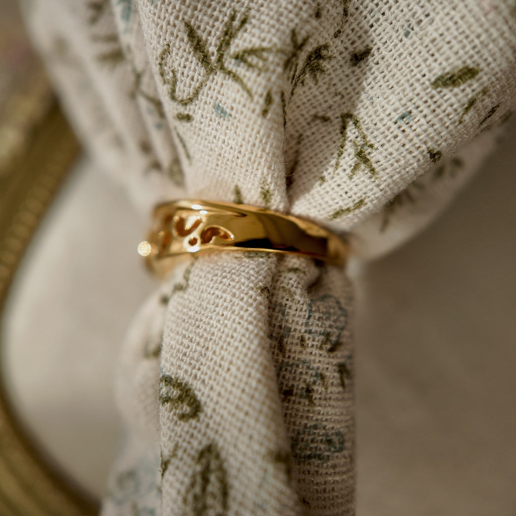 The 23k gold-plated ring features a round glass diamond and an openwork design with intricate patterns. The vintage ring, worn through a floral fabric weave, reveals from the side that the design fades towards the back, leaving only a plain gold band.