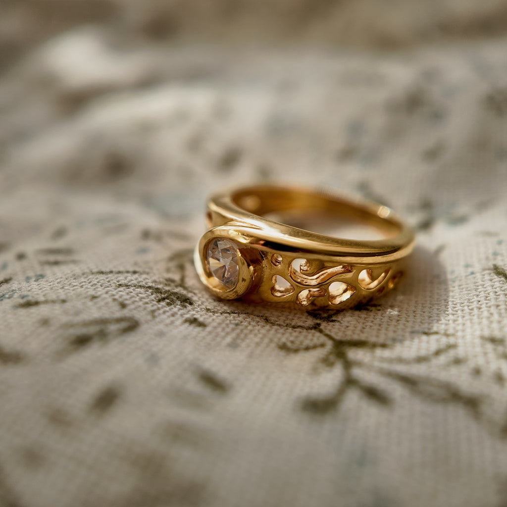 The 23k gold-plated ring features a round glass diamond, and the band is openwork-carved with intricate patterns. The vintage ring is placed on a floral fabric; from a side view, different openwork patterns are visible on the side of the ring.