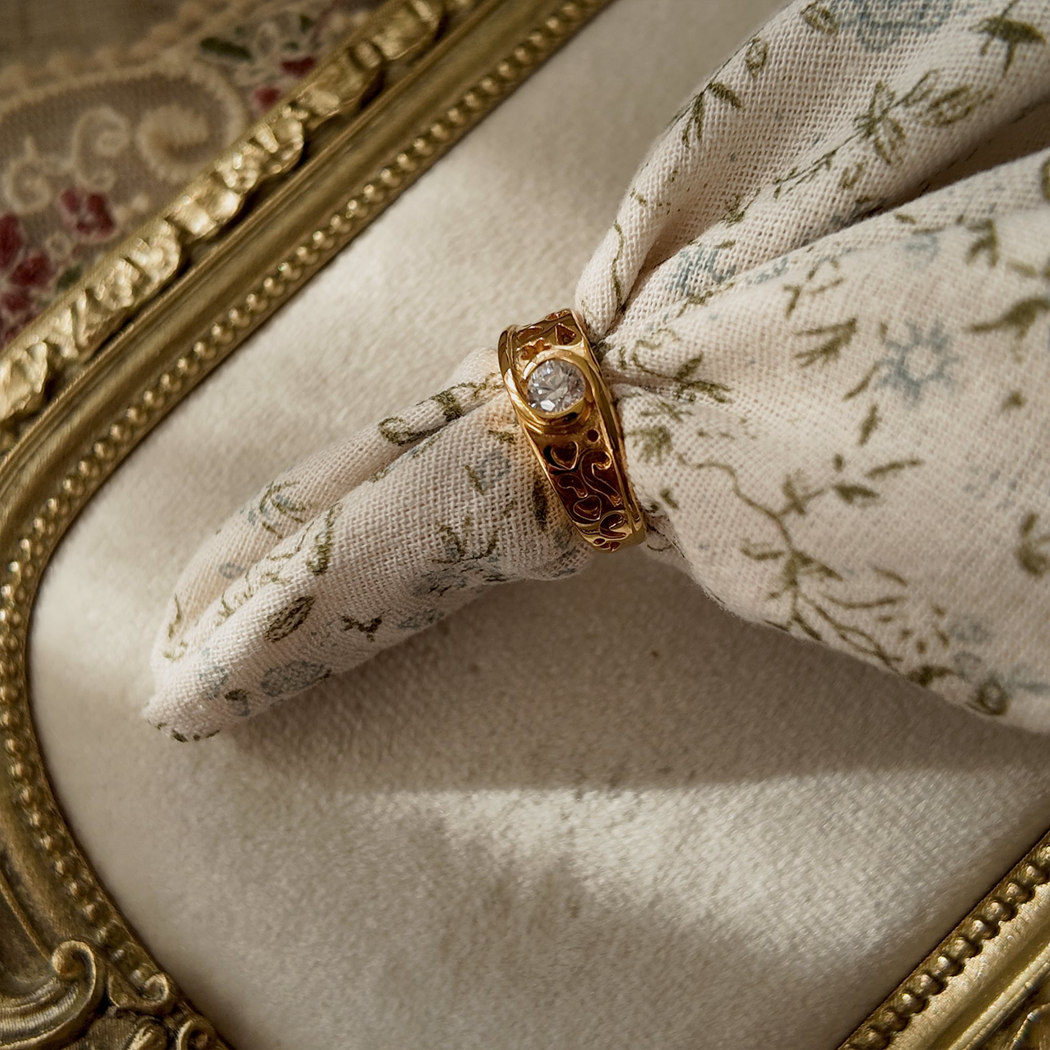 The 23k gold-plated ring features a round brilliant-cut glass diamond, and the band is openwork-carved with delicate patterns. The vintage ring is strung with floral fabric to simulate how it would look on a hand. The diamond is lustrous, and the band features cute patterns resembling auspicious clouds and hearts.