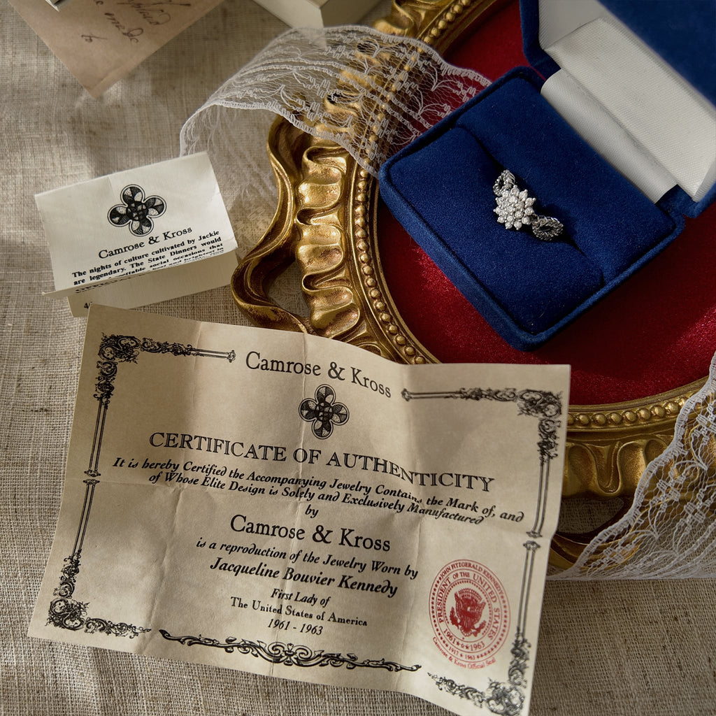 The silver ring, adorned with glass diamonds arranged in a floral pattern, is a collaboration between Camrose & Kross and JBK, and is presented in a ring box. A magnified view of the authentication card is also included.