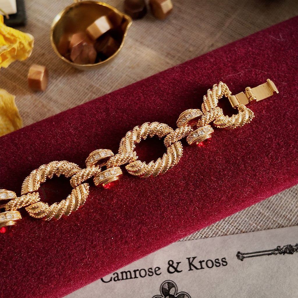 The gold-brushed bracelet with red gemstones sits in its original red velvet box, highlighting the clasp details.