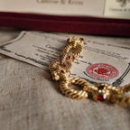 The gold bracelet is placed on the official certificate under sunlight; a close-up side view shows the red gemstone at the joint.