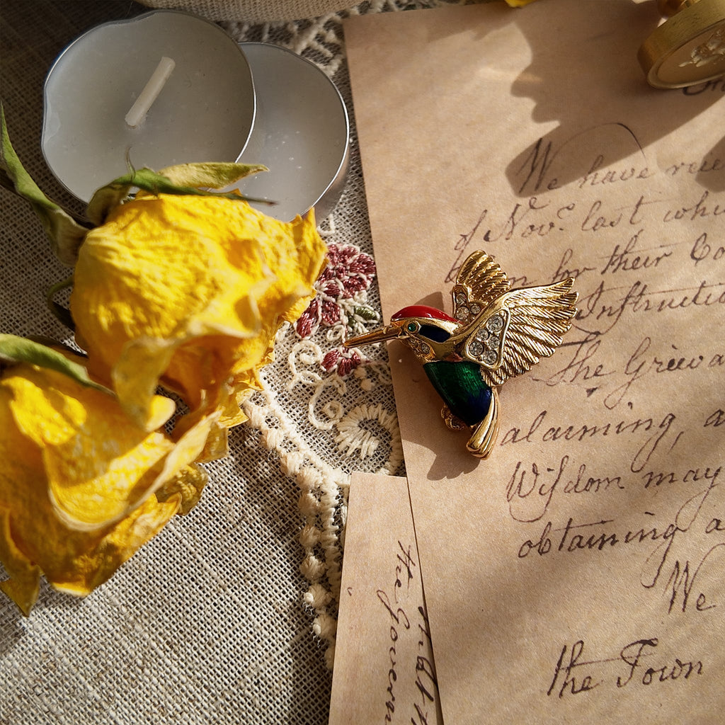 A brooch featuring a hummingbird design, with a head made of red enamel, a face of black enamel, and a body of blue and green enamel. The hummingbird's chin and wings are adorned with small rhinestones, and the entire piece is gilded. The wings are decorated with carved feather patterns. The eyes are set with a green gemstone, giving it a bright and expressive look.