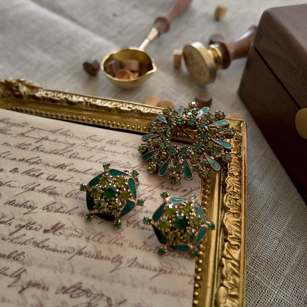 A vintage enamel brooch set with an emerald green ring of rhinestones resembles a bursting firework, and the set also includes a pair of round clip-on earrings. Both pieces are composed of toned enamel and high-quality rhinestones and are from the brand WEISS.