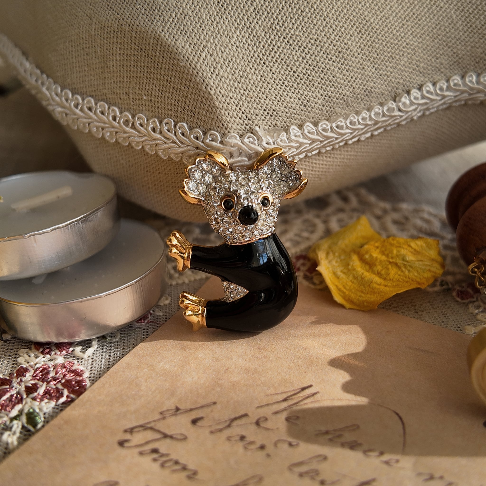 This adorable koala brooch sits sideways on a jewelry stand. The koala's face and ears are adorned with several sparkling mini rhinestones, and the entire piece is edged in gold. The body is filled with black enamel, while the hands and feet remain gold, with a few rhinestones added to the belly. The koala has a cute, slightly dazed expression and a posture resembling that of someone hugging a tree trunk. This incredibly cute koala brooch is remarkably lifelike.