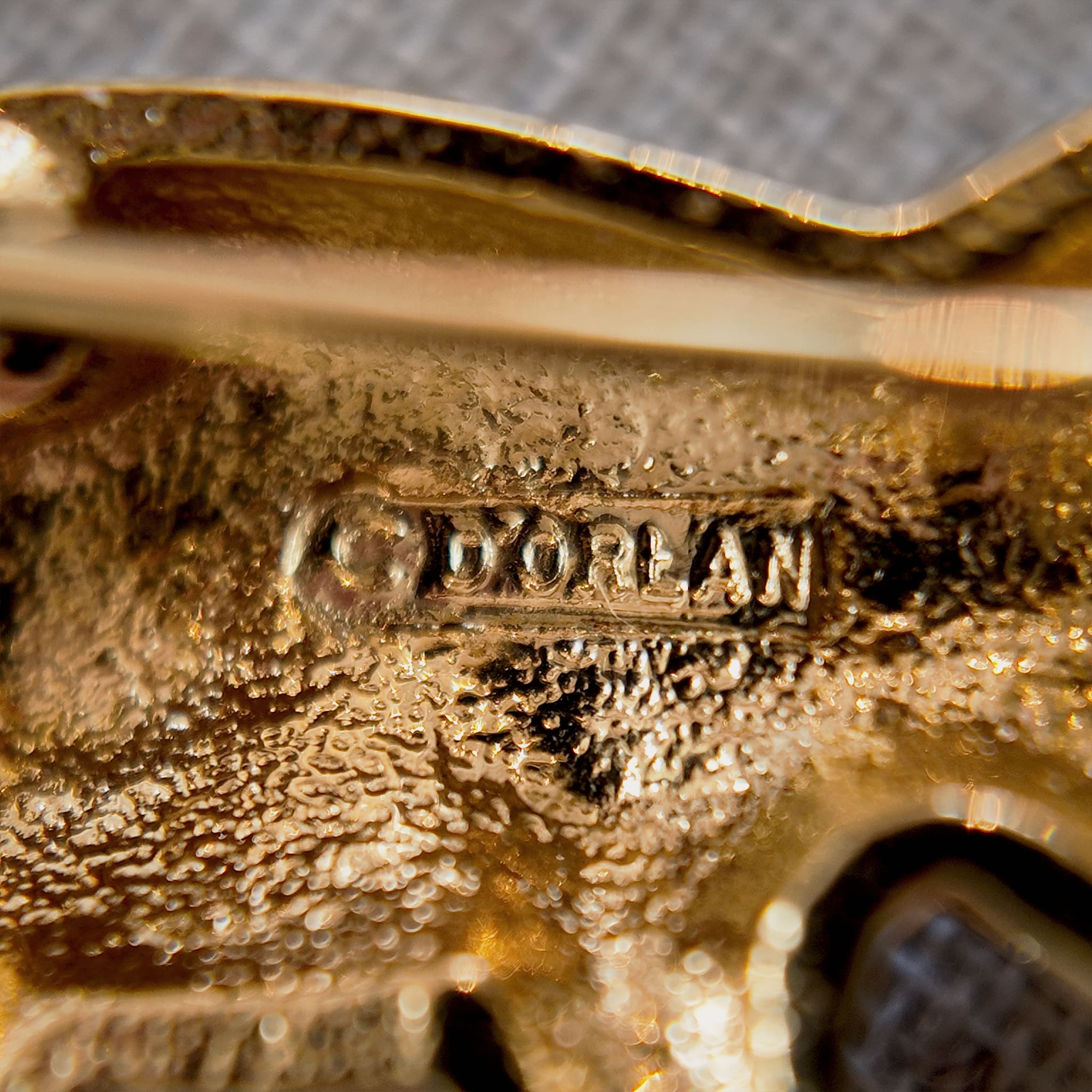 This koala brooch from D'orlan lies in the sunlight, and a close-up of the brand's embossed logo on the back allows for a clearer view and identification of the brand's provenance.