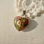 A bronze kitten head sits atop a heart-shaped natural stone pendant. The stone is a combination of seaweed green and coral pink, with irregularly stained natural stone. Heart-shaped stone pendant.