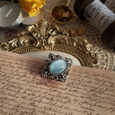 A pale blue imitation moonstone is set in a silver brooch with Victorian-style carvings. This vintage brooch rests sideways on a jewelry stand, showcasing its front view.