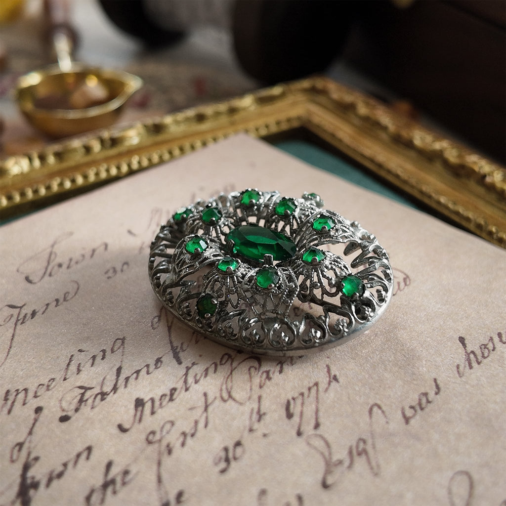 The silver cloisonné brooch is set with an emerald gemstone. A close-up side view highlights the texture of the central oval emerald gemstone.