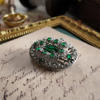 The silver cloisonné brooch is set with an emerald gemstone. A close-up side view highlights the texture of the central oval emerald gemstone.