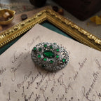 A silver cloisonné brooch is set with emerald gemstones. A medium-shot side view with a zoom-in reveals the texture of the small emerald gemstones set around the vintage brooch from a side angle.