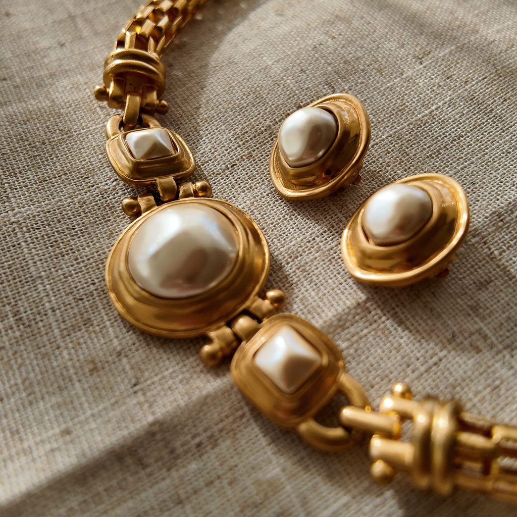 A pair of vintage Anne Klein clip-on earrings, featuring a central baroque pearl surrounded by a bold, circular frame with a rich, brushed matte gold finish.
