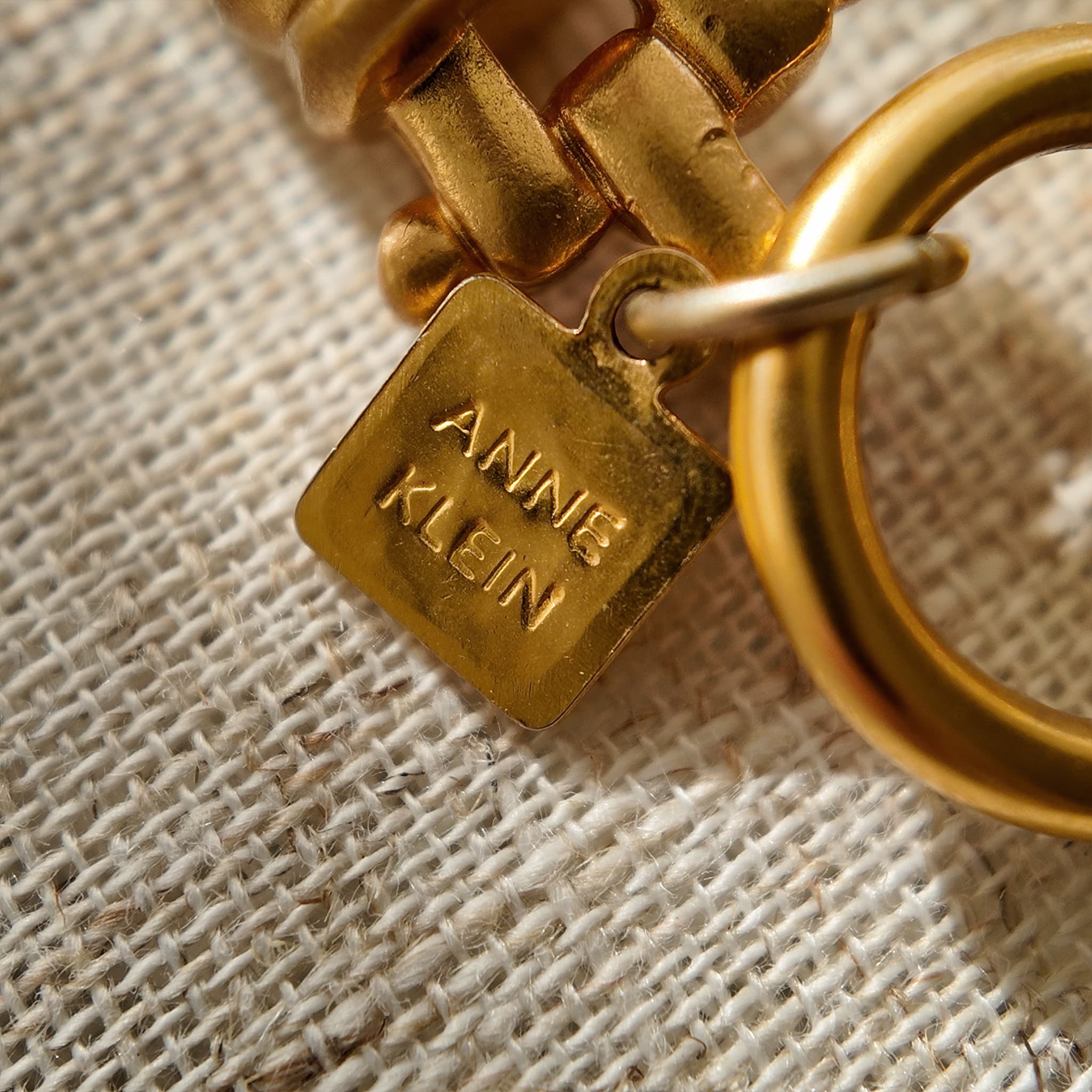 A detailed photo of the authentic Anne Klein signatures: the iconic lion head logo hang tag on the necklace and the signature "AK" hammered texture on the reverse.