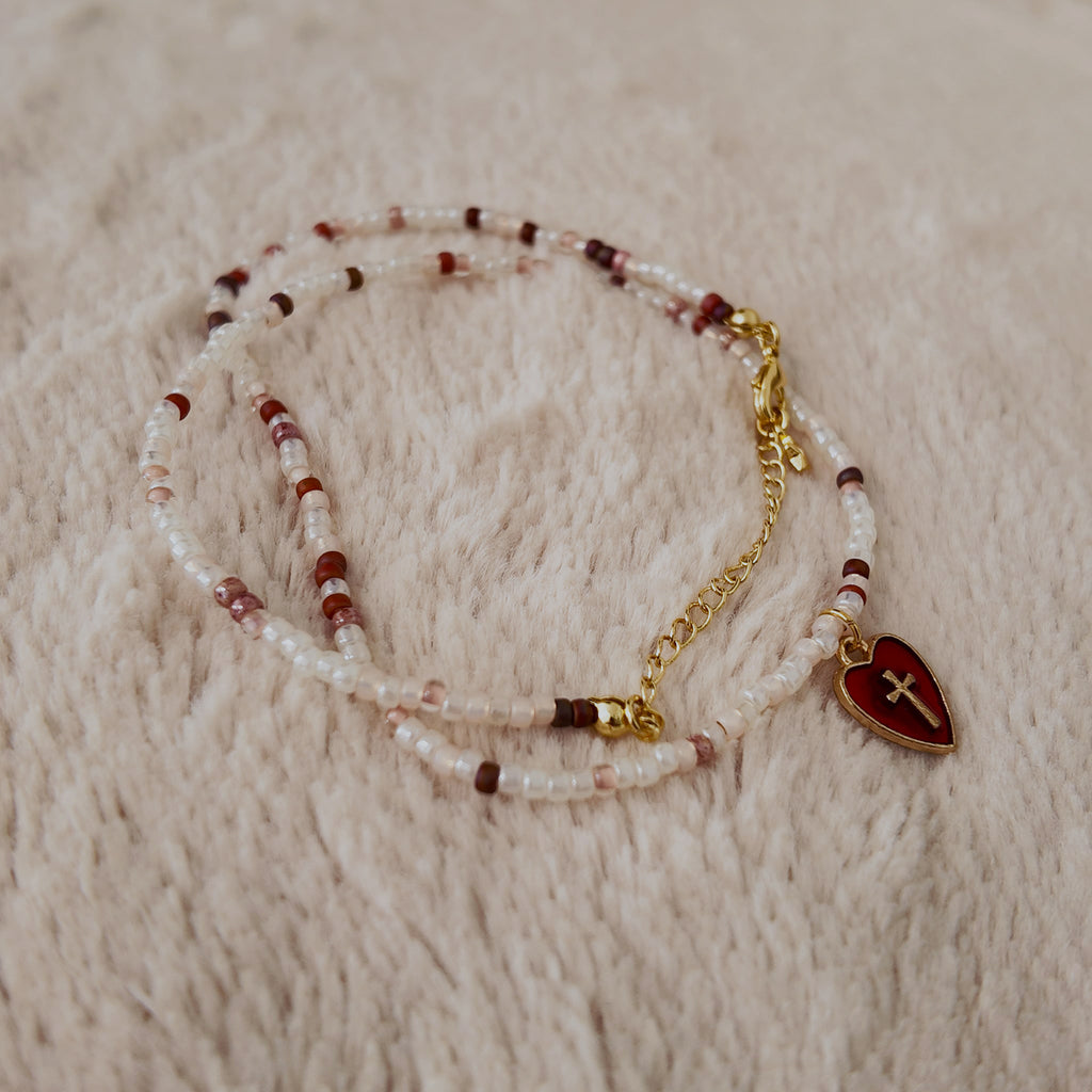 An angled view of the handmade necklace, highlighting the bold, modern anchor of the red metal Heart-Cross charm against the delicate, rhythmic beading.