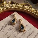 An angled view of the sapphire blue earrings, highlighting the delicate, lace-like filigree work that frames the central teardrop and smaller clear crystals.