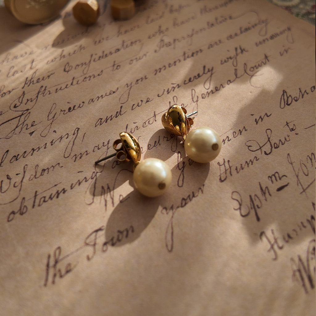 An angled view of the gold dangle earrings, highlighting the playful movement of the luminous pearl suspended below the polished heart stud.