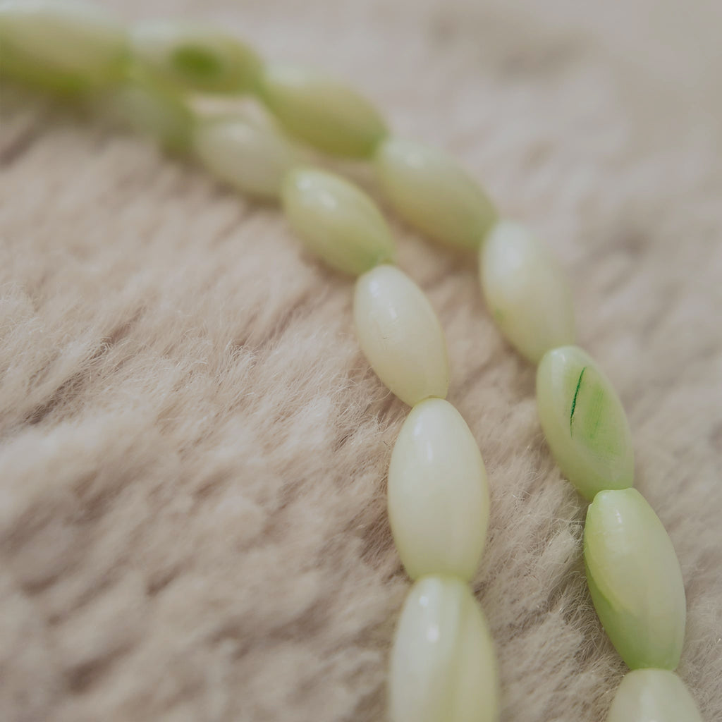 A macro shot detailing the sleek, tapered shuttle shape of the natural shell beads and the unique, soft blend of milky white and mint green colors.