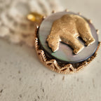 A close-up of a mythological bear pendant, showing the detailed vintage brass bear seemingly walking across a night sky of reflective Black Mother-of-Pearl.